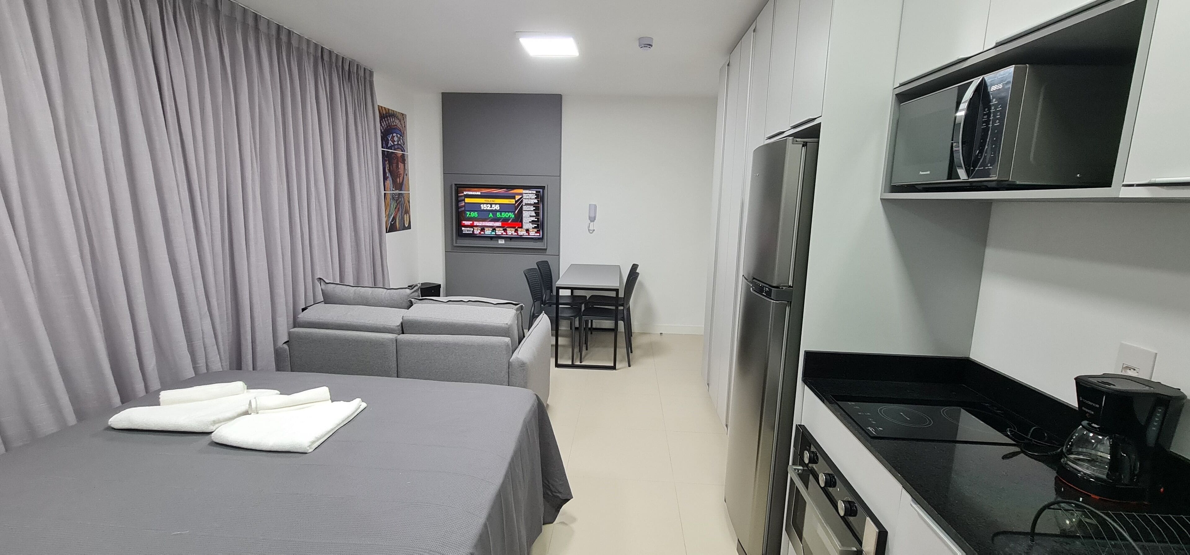 A compact hotel room with a bed, a sofa, a small dining table, a kitchen area with a refrigerator and stove, and a TV mounted on the wall.