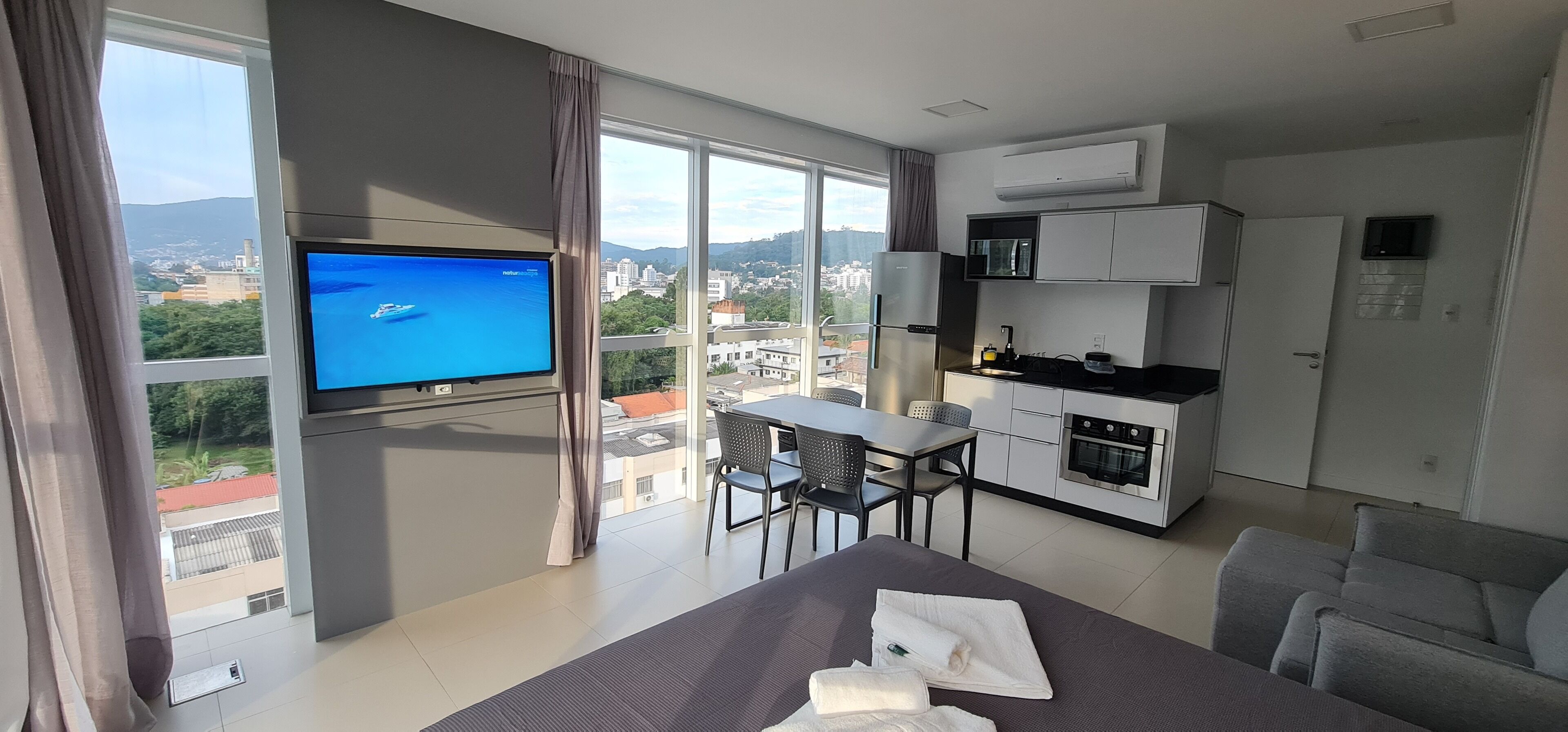 Una camera d'albergo moderna con una grande finestra, una televisione a schermo piatto, un'area pranzo e un angolo cottura.
