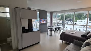A modern hotel room with a kitchenette, a dining area, a bed with a view, and large windows.