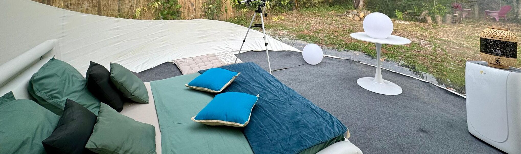 Area esterna coperta da tende, con un letto, cuscini, un tavolo e un oggetto sferico, più una lanterna.