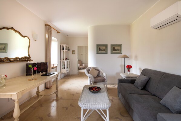 A living room with a grey sofa, a white wicker coffee table, a dining table with a vase of flowers, and a white armchair. - Dependance a Baglio il Corvo by Wonderful Italy (Geraci Siculo)