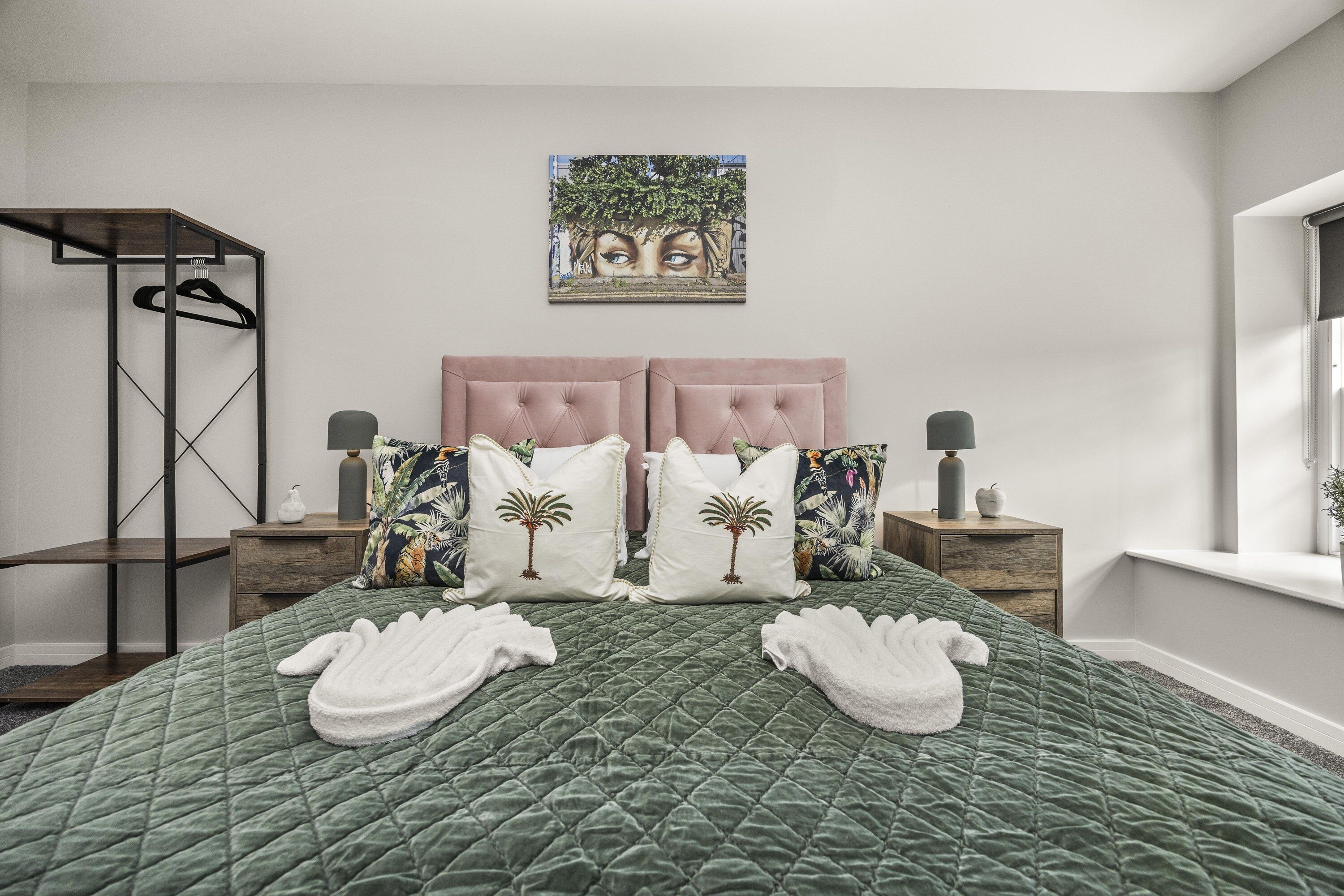 A neatly made bed with a green quilt, two nightstands with lamps, a wooden side table, a wall-mounted artwork, and a window with a view.