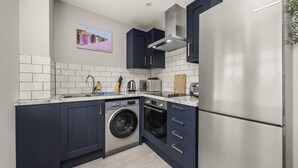 A compact kitchen with dark cabinets, a stainless steel refrigerator, a washing machine, and a toaster. - Buzzing Brighton Apartment 6 | By My Getaways (Brighton)