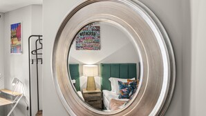 A bedroom with a green upholstered headboard, a bedside table with a lamp, and a mirror reflecting the room. - Buzzing Brighton Apartment 6 | By My Getaways (Brighton)