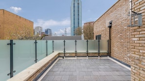 A rooftop terrace with a glass balustrade, brick walls, and a view of a tall building in the background. - Guest Ready - Oak Crescent City House, London, E14 (London)