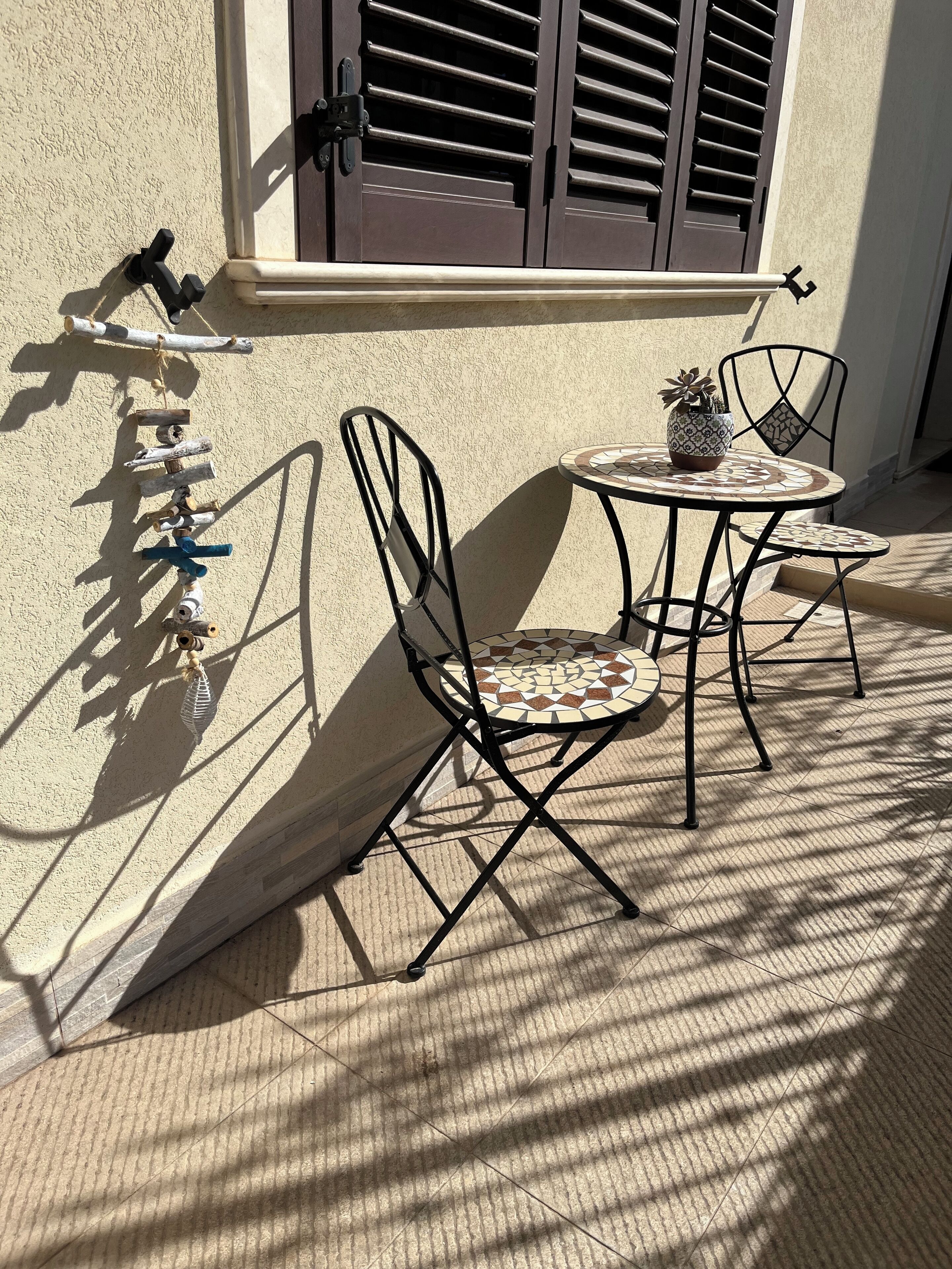 Ein kleiner Sitzbereich im Freien mit zwei Metallstühlen und einem Tisch, einem Topfpflanzen und einem dekorativen Metallregal an einer Wand.