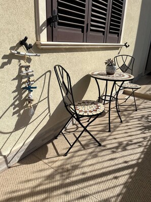 Un petit espace extérieur aménagé avec deux chaises en métal et une table, un pot de fleurs et un présentoir métallique décoratif adossé à un mur.