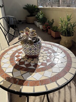 Une table ronde ornée d’un motif en mosaïque, entourée de plantes en pot, et une chaise en métal.