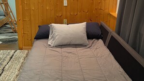 Un dormitori amb una paret de fusta panelada, dues làmpades penjades a la paret, un llit amb edredó i almoines grises, una petita taula de nit i una finestra amb cortines.