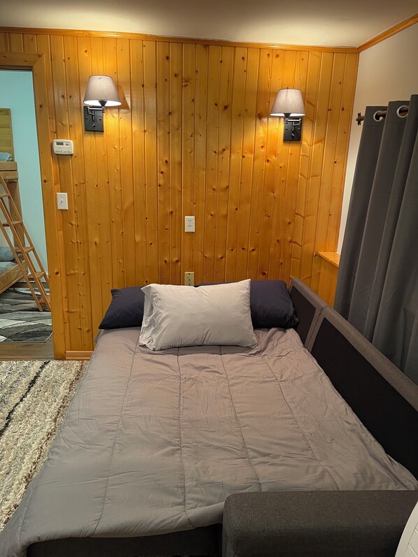 Une chambre avec un mur lambrissé en bois, deux lampes fixées au mur, un lit avec une couvre-lit et des oreillers gris, une petite table de chevet et une fenêtre avec des rideaux.