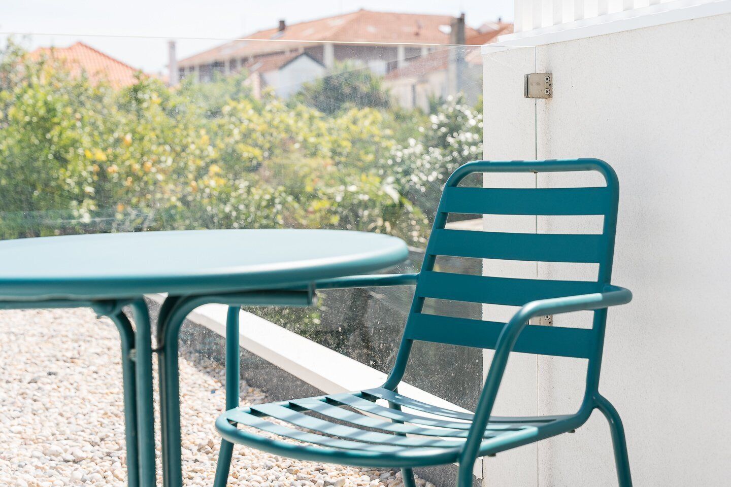 Een set van een teal metalen stoel en tafel voor een witte muur, met zicht op groen en gebouwen op de achtergrond.