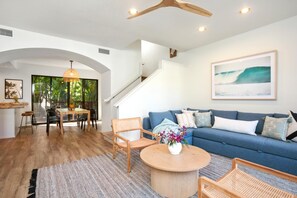 Ein modernes Wohnzimmer mit einem blauen Sofa, einem Holztisch und Korbstühlen. Eine Treppe führt zu einer weiteren Ebene, und im Hintergrund ist ein Essbereich zu sehen.