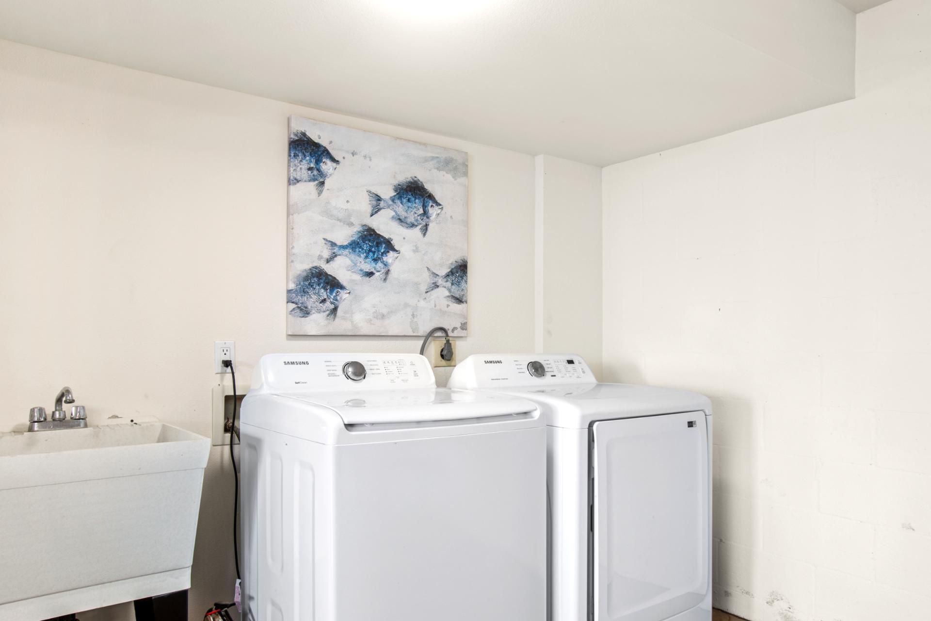 Un espace buanderie comprenant deux machines à laver Samsung blanches, un évier blanc et un tableau représentant des poissons, accroché au mur.