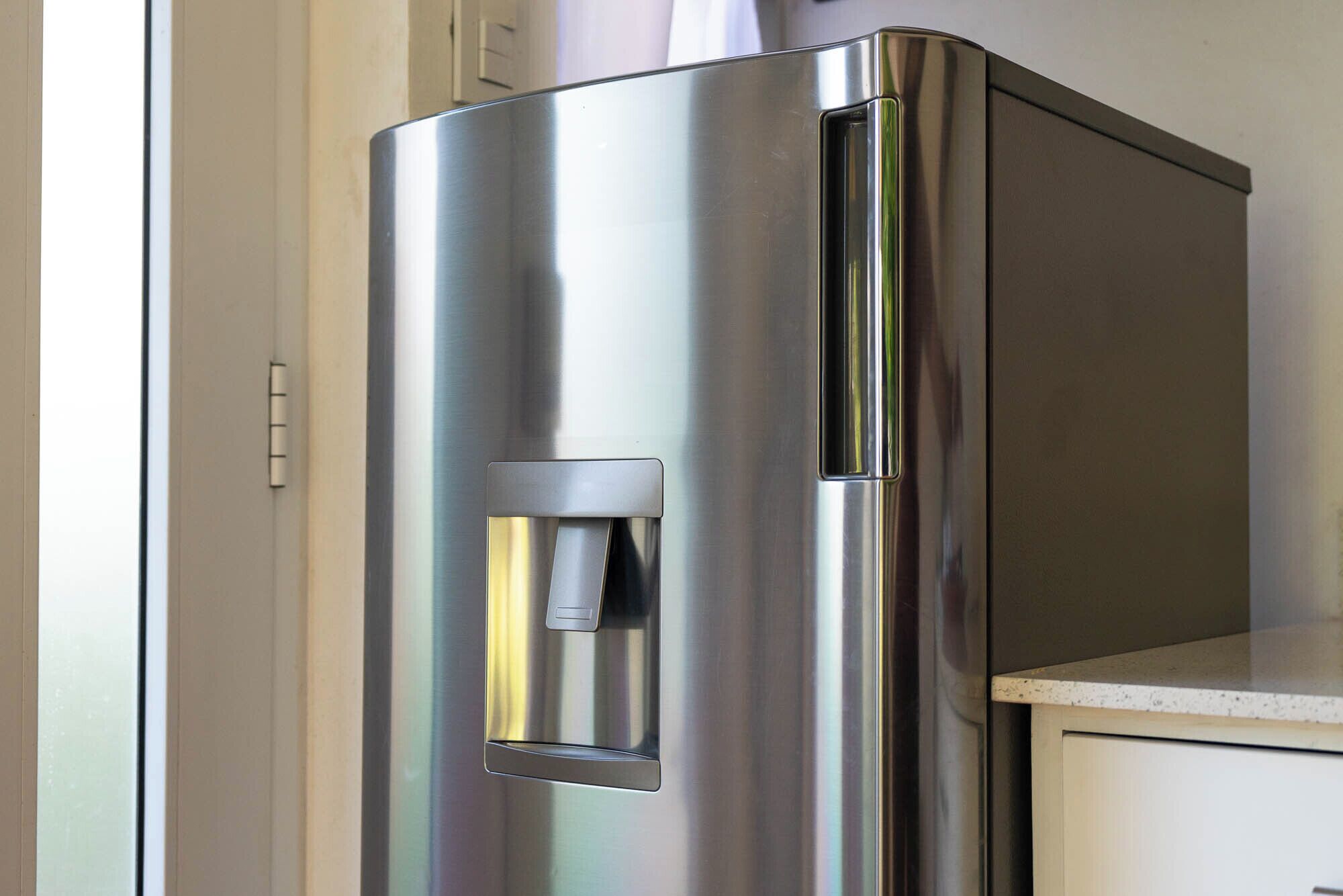 A stainless steel refrigerator with a water dispenser, situated in a kitchen with a white countertop and a cabinet.