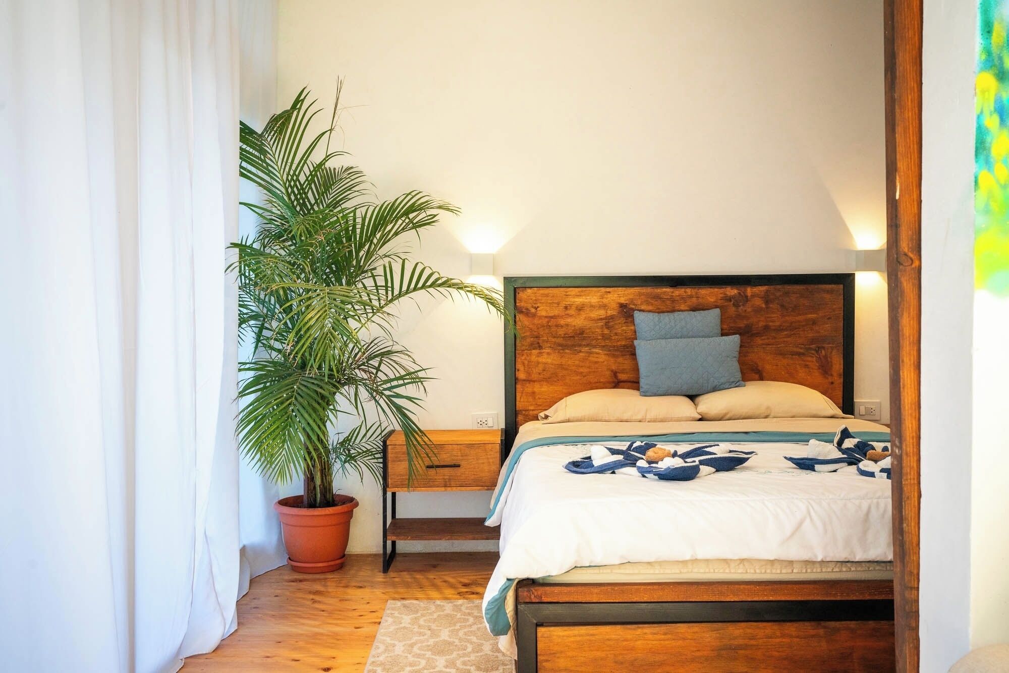A bedroom with a wooden bed, a nightstand, a potted palm tree, and a window with curtains.