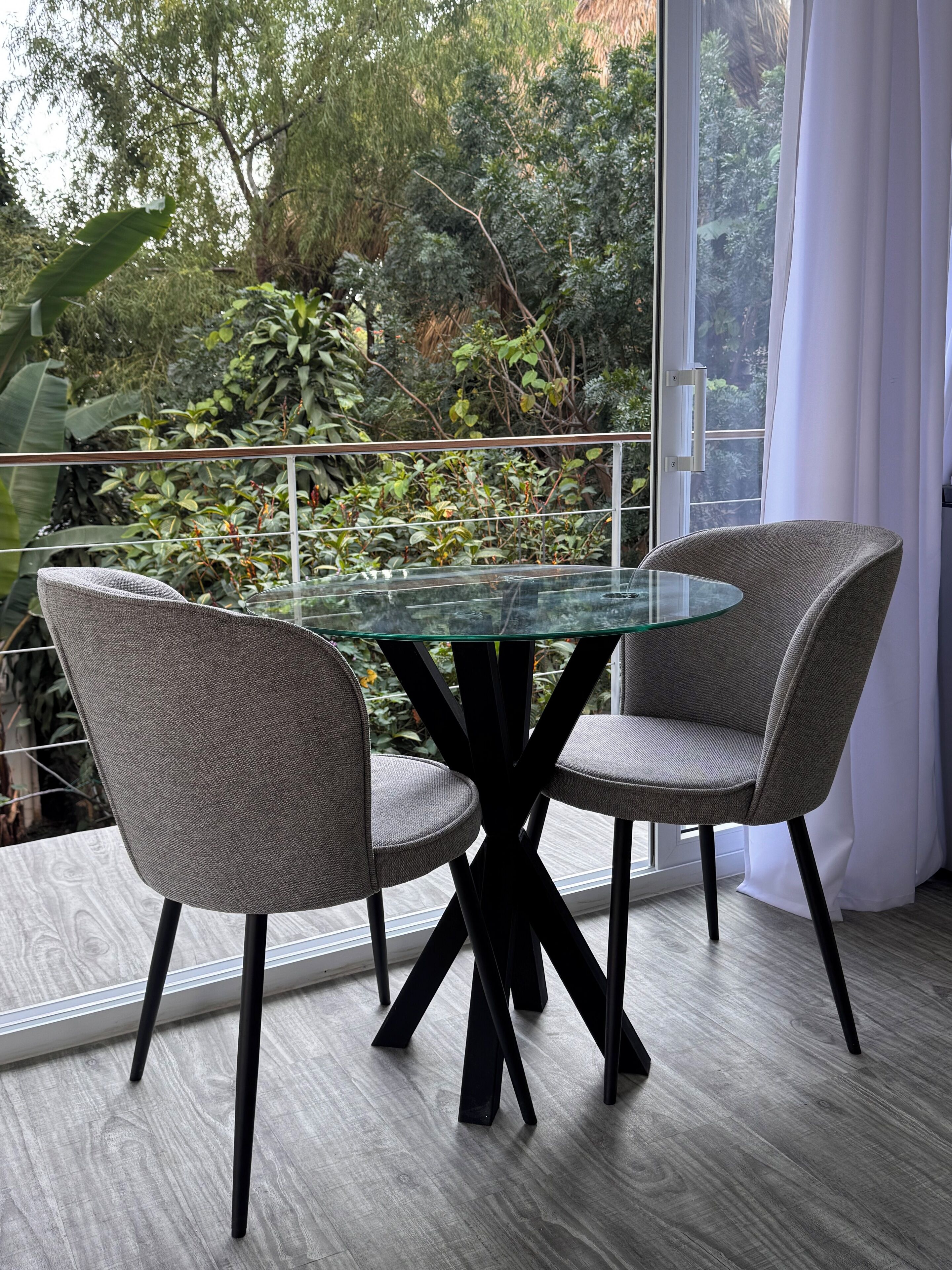 Un espace repas moderne avec une table en verre et deux chaises grises à revêtement, le tout mis en valeur par un décor de verdure luxuriante et une grande fenêtre agrémentée de rideaux blancs.