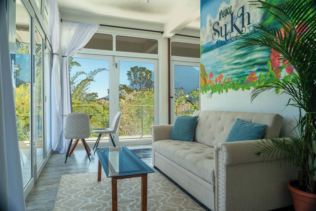 Una sala de estar moderna con un sofá color beige, dos sillones, una mesa de centro de vidrio y un ventanal con vistas a zonas verdes y montañas.