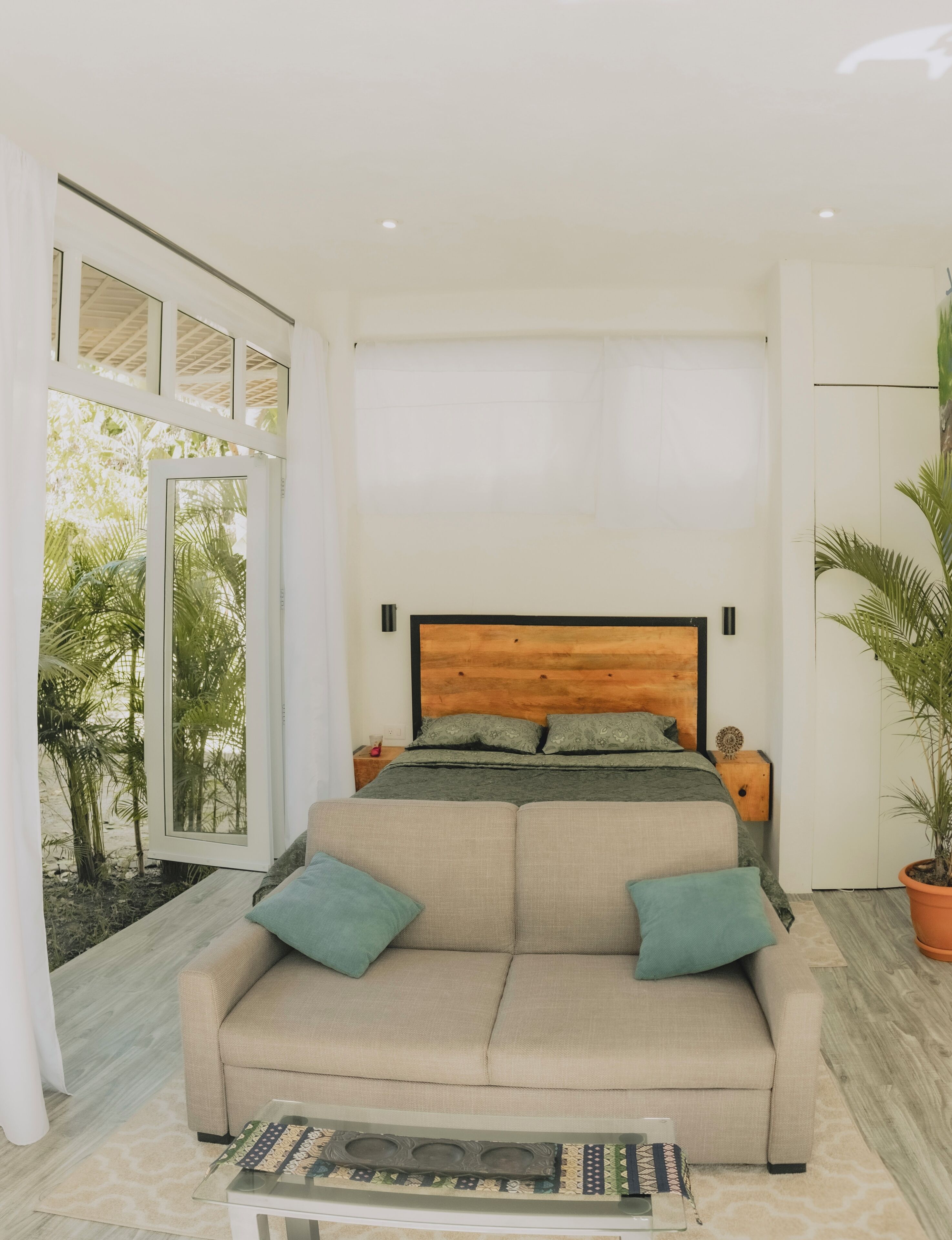 Une chambre avec une tête de lit en bois, un canapé avec des coussins, une table basse en verre et une vue sur la verdure à l’extérieur.