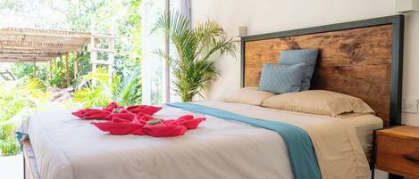 A neatly made bed with a wooden headboard, a bedside table, and a view of lush greenery outside.