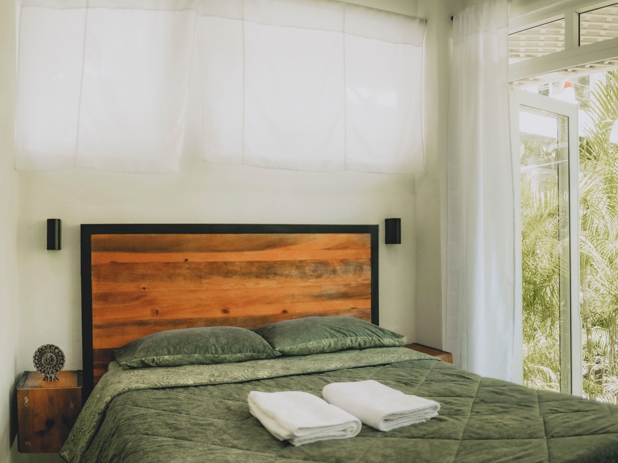 A bedroom with a wooden headboard, green bedding, and a view of greenery through a window.