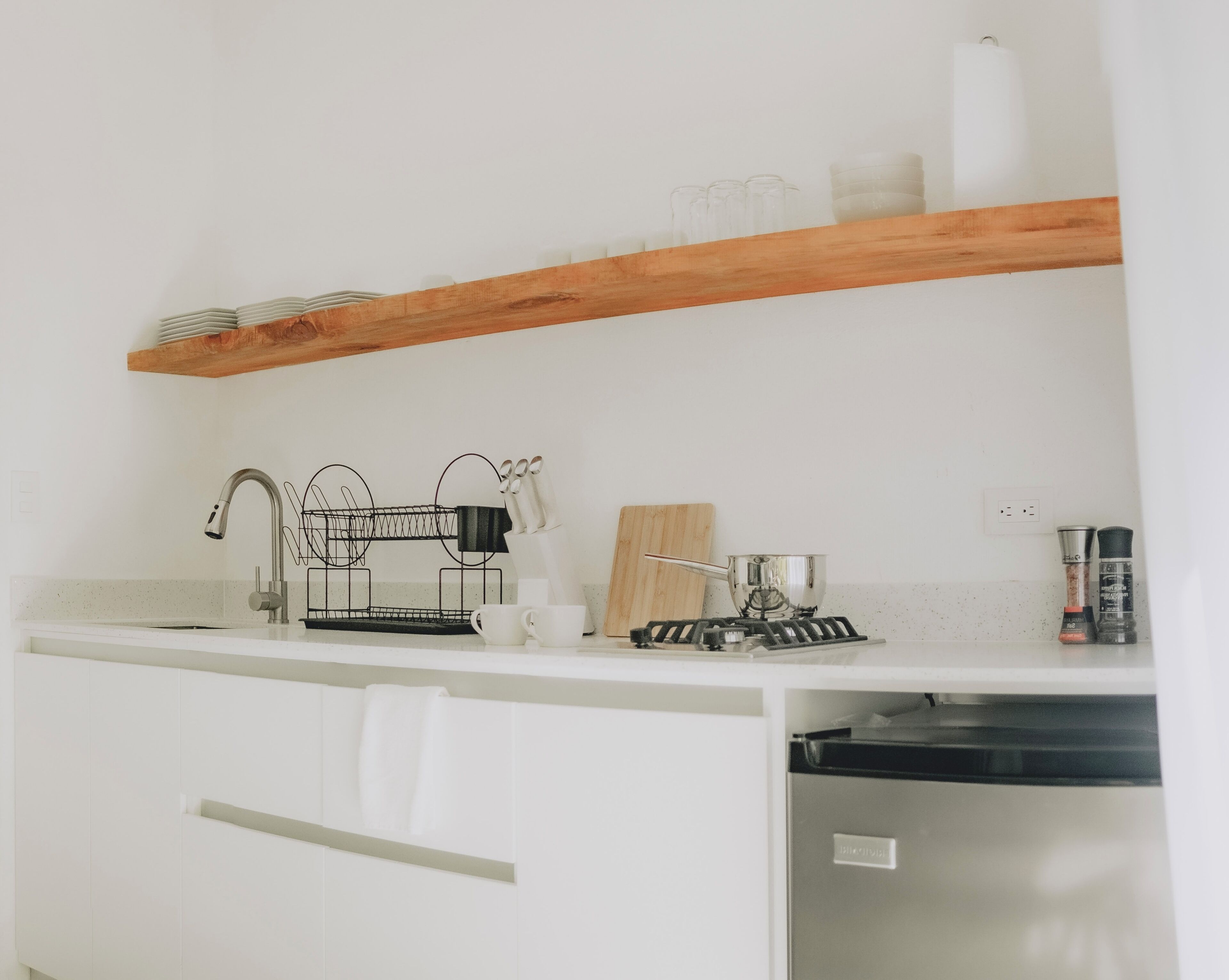 A kitchen with white cabinets, a wooden shelf above the counter, a coffee maker, a kettle, and a dish rack.