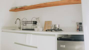 A kitchen with white cabinets, a wooden shelf above the counter, a coffee maker, a kettle, and a dish rack.