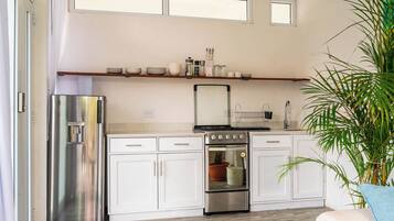 A modern kitchen with white cabinets, a stainless steel refrigerator, a gas stove, and a built-in oven. There is a window above the sink and a shelf with kitchenware.