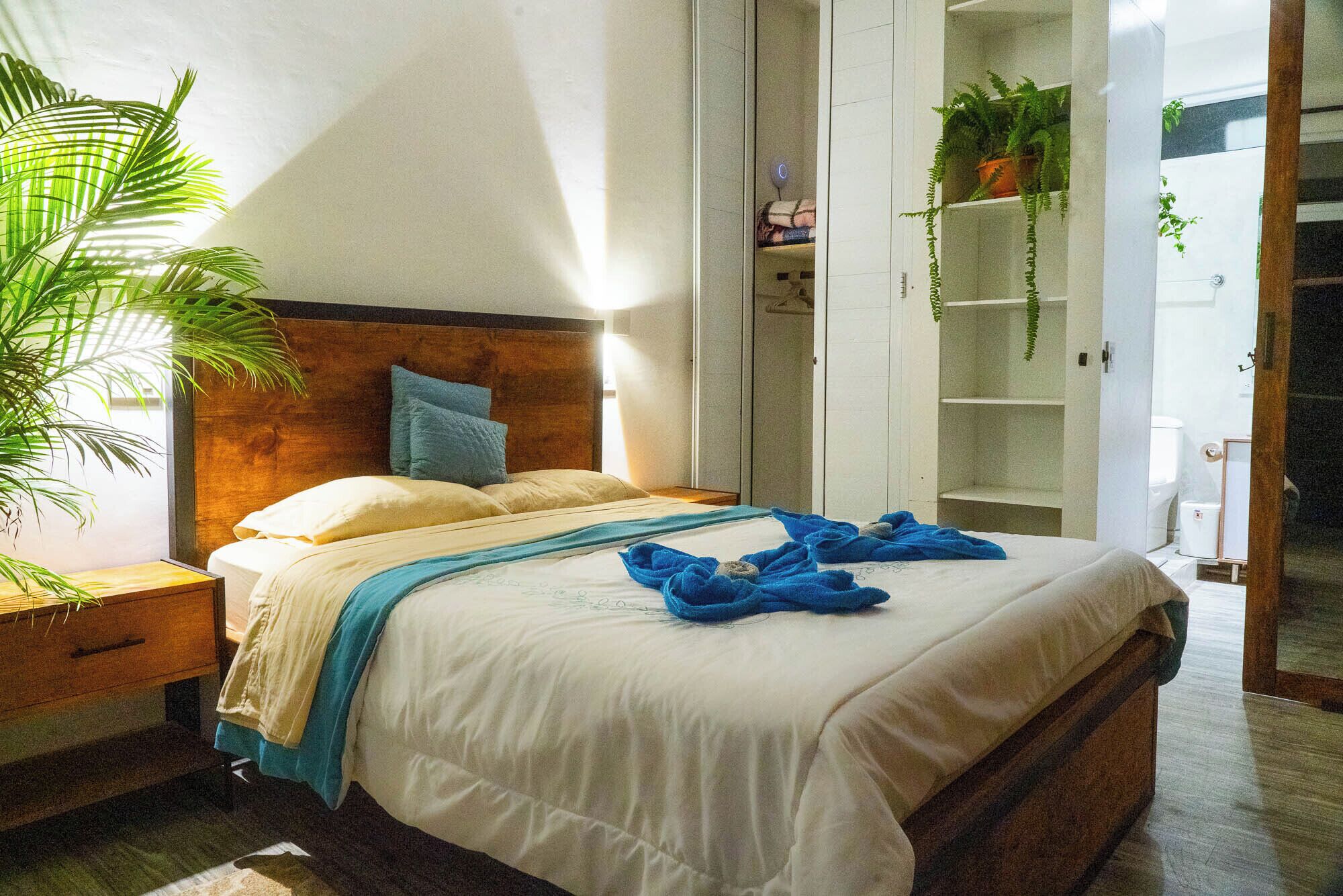 Een net opgemaakt bed met een houten hoofdbord, een nachtkastje en een grote potplant aan de linkerzijde.