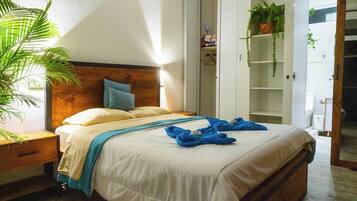Een net opgemaakt bed met een houten hoofdbord, een nachtkastje en een grote potplant aan de linkerzijde.