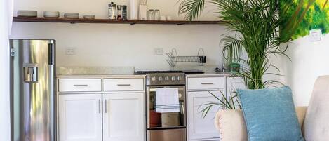Une cuisine compacte avec des meubles blancs, un réfrigérateur en acier inoxydable, une cuisinière et un lave-vaisselle. Une étagère en bois se trouve au-dessus du comptoir, où sont rangés des ustensiles de cuisine et une plante en pot. Un canapé beige avec un coussin bleu est visible au premier plan.