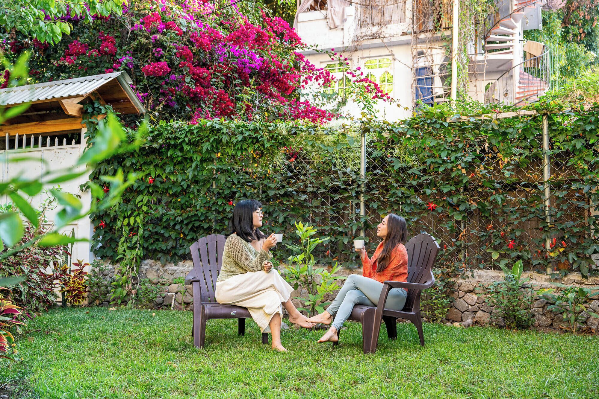 Two people seated on wooden chairs in a garden with a stone wall and flowering plants in the background.