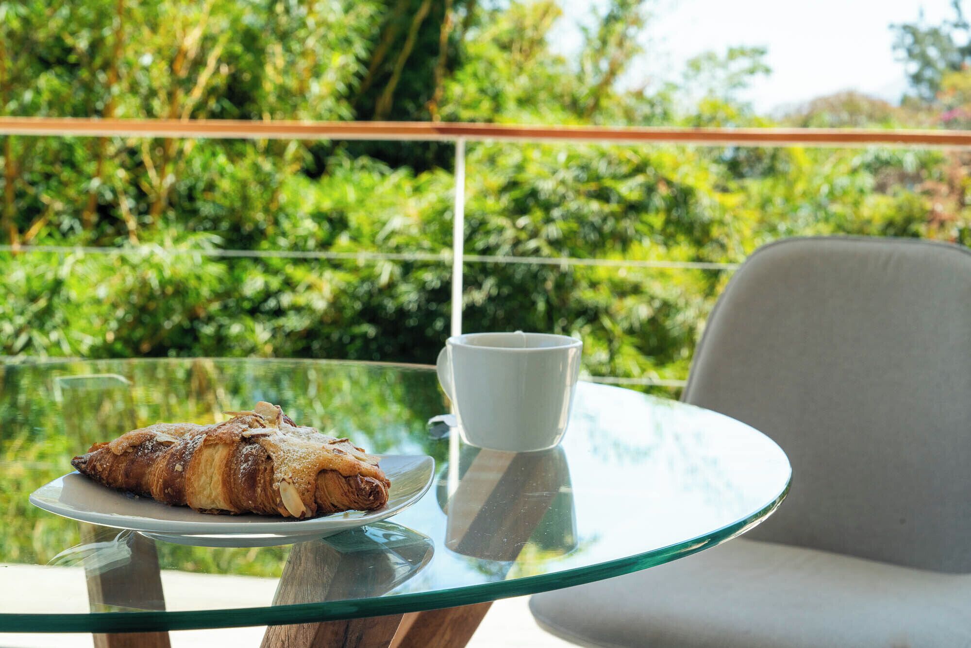 Una mesa de vidrio con un pastelito y una taza, sobre un fondo verde exuberante.