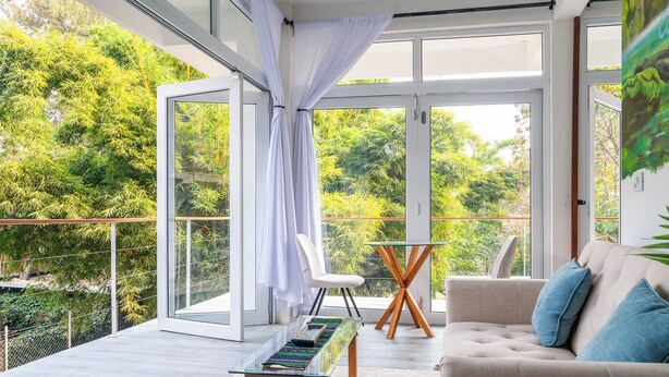 Un salón moderno con una mesa de vidrio, un sofá y una silla, con balcón y ventanas amplias con vistas a zonas verdes.