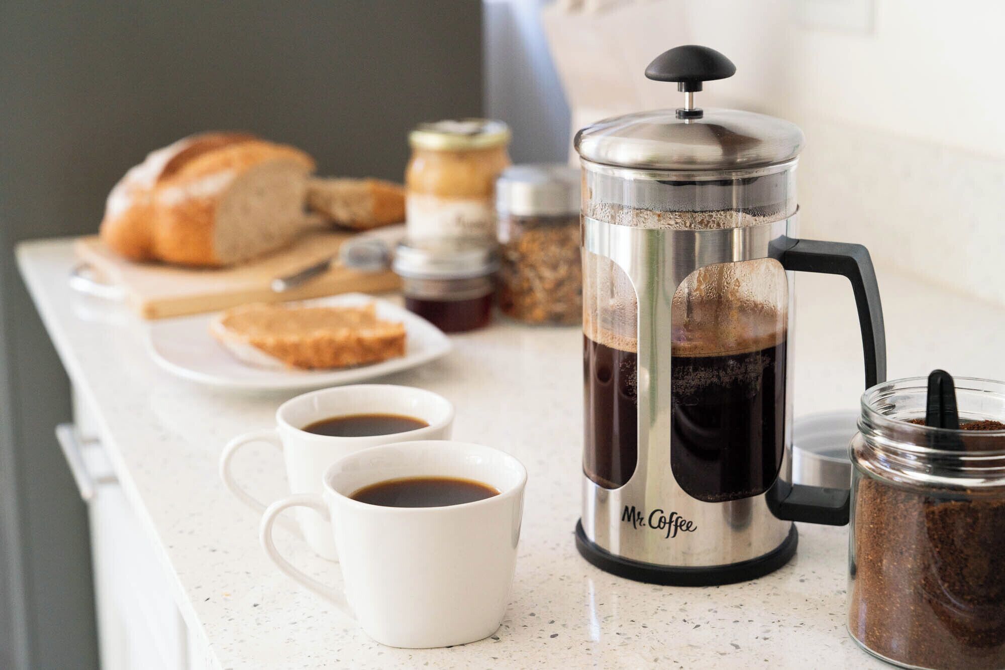 Une cafetière à piston, de la marque « Mr. Coffee », est posée sur un plan de travail de cuisine, à côté de deux tasses de café, d’un bocal de grains de café, d’une miche de pain et d’un pot de miel.