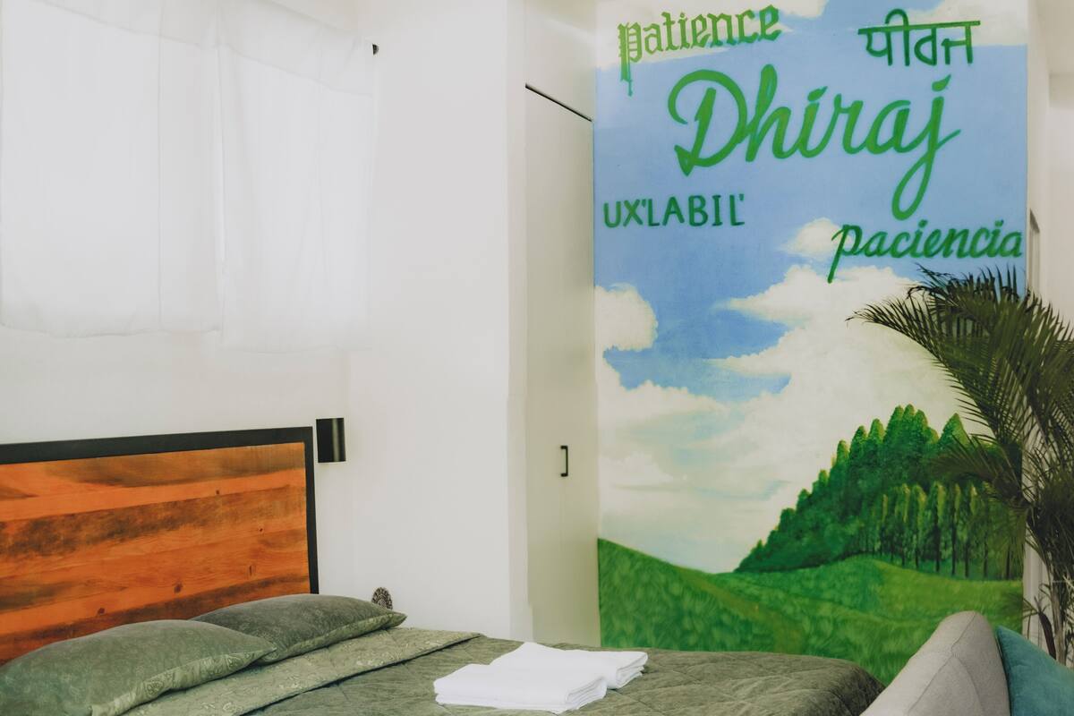 Un dormitorio con cama, mesita de noche, pared con un mural de temática natural y una puerta con texto en varios idiomas.
