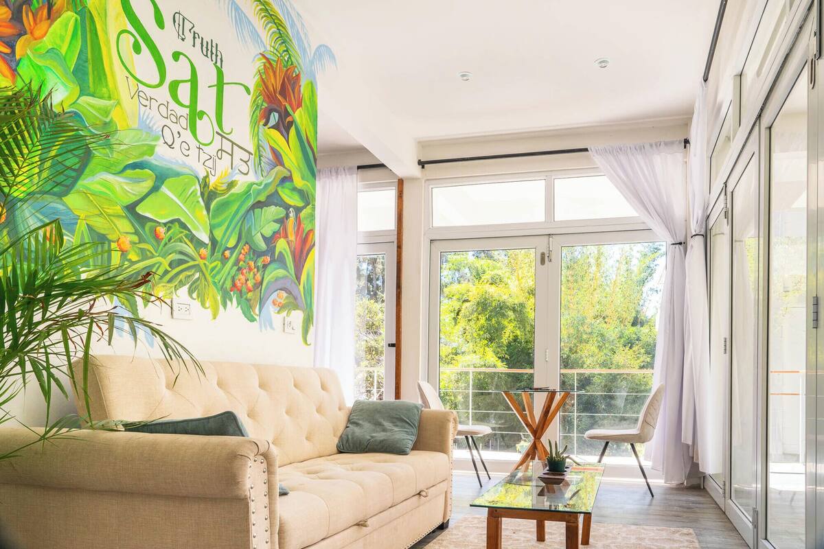 Una sala de estar moderna con un mural grande, un sofá color beige, una mesa de centro de vidrio y vista a zonas verdes al exterior.