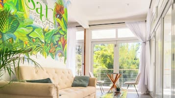 Een moderne woonkamer met een groot muurschildering, een beige bank, een glazen salontafel en uitzicht op groen.