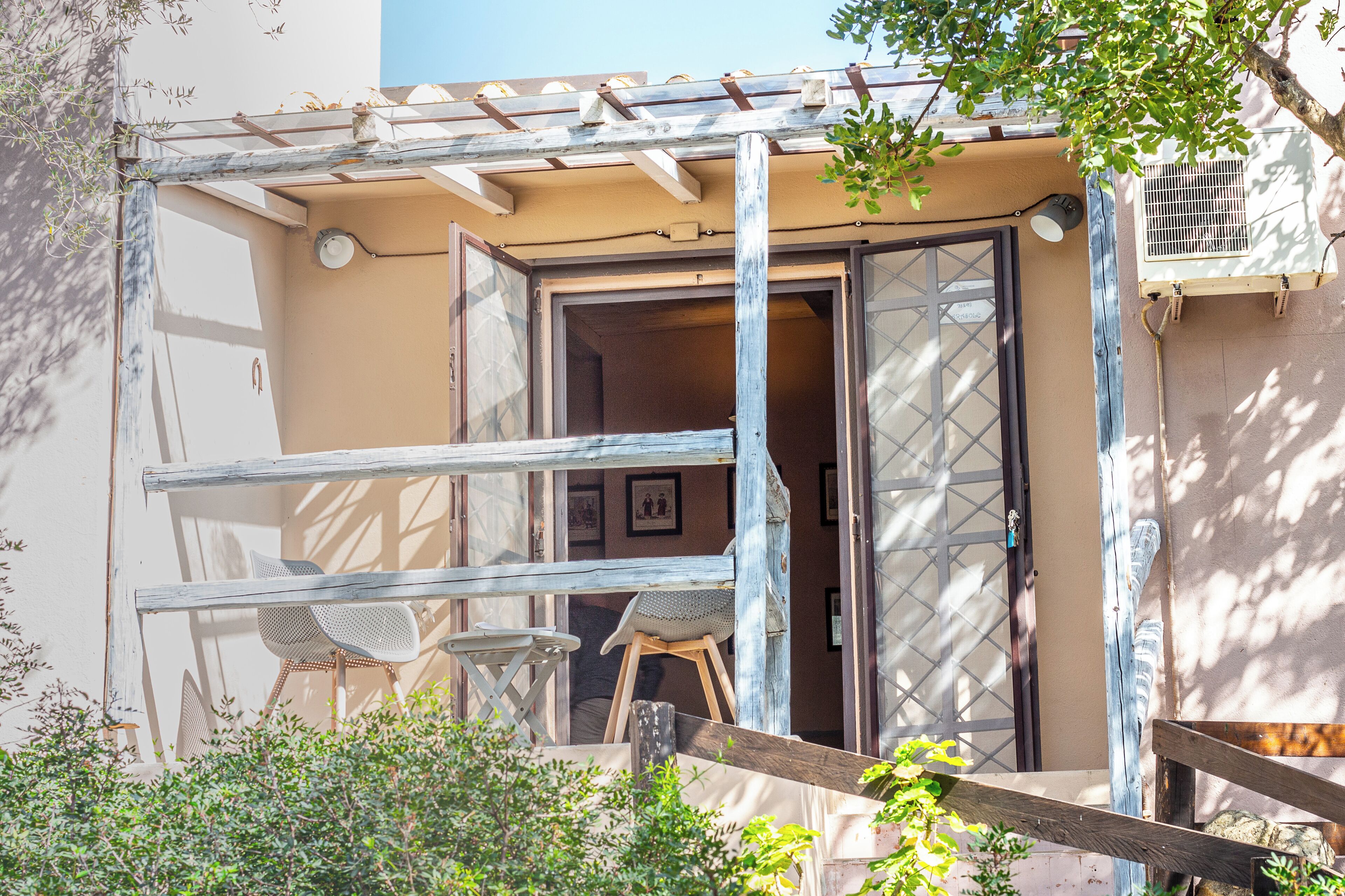 Een gebouw met een houten pergola, een deur met een ruit en een kleine tafel met stoelen buiten.