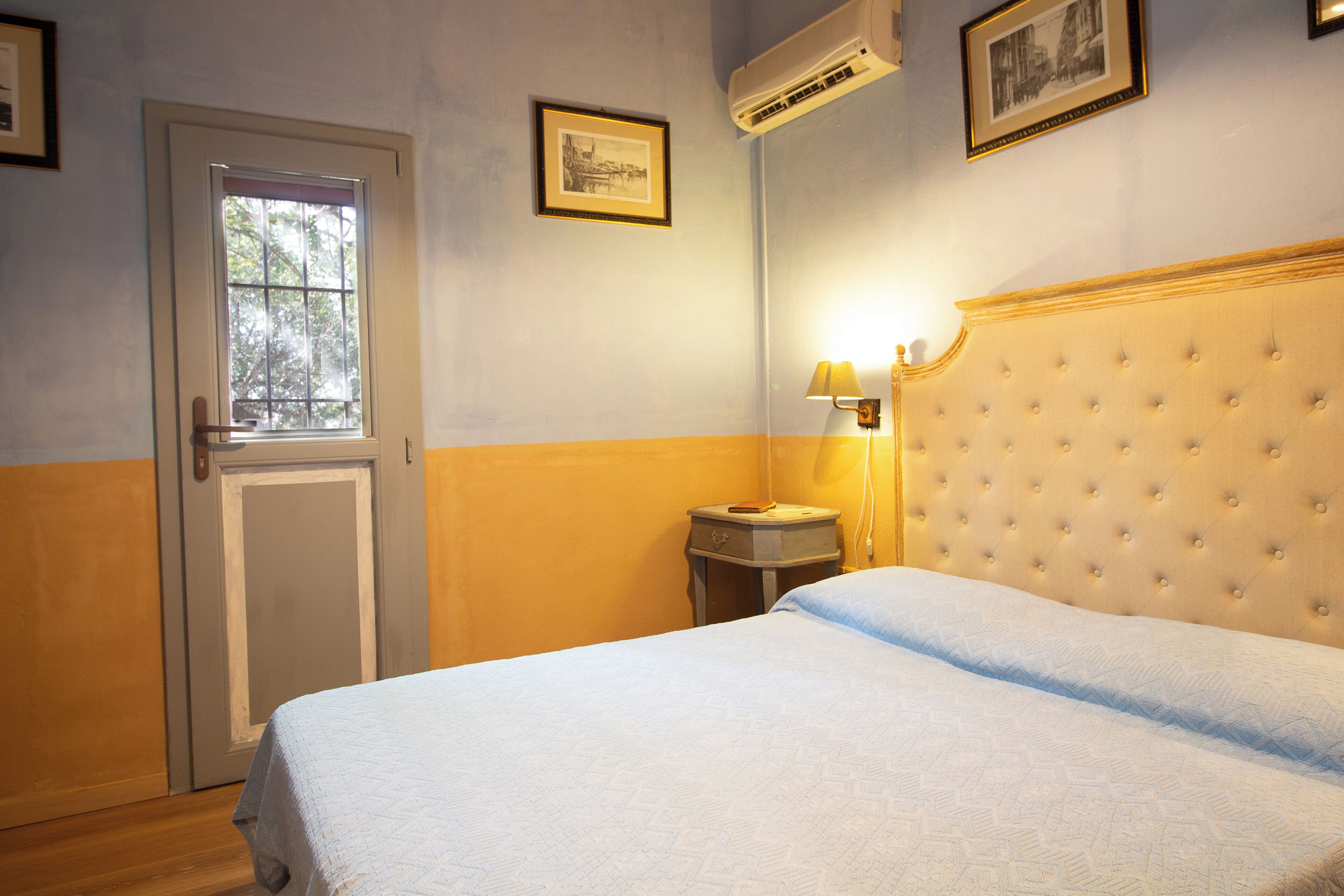 A bedroom with a bed, a nightstand, a lamp, a door, and an air conditioning unit.