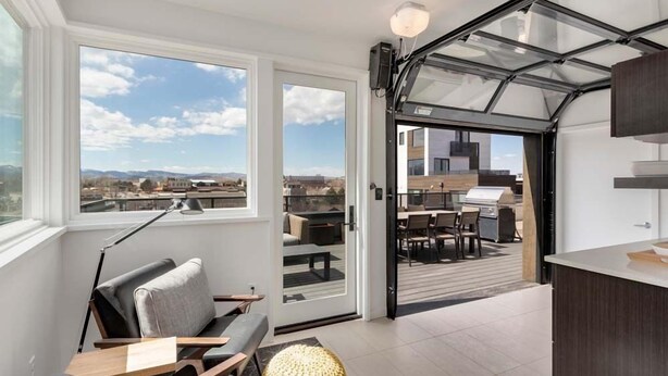 A modern interior with a view of the cityscape through large windows, a sliding glass door leading to an outdoor patio, and a kitchen area.