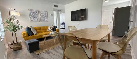 A modern living room with a wooden dining table, yellow sofa, and a TV mounted on the wall.
