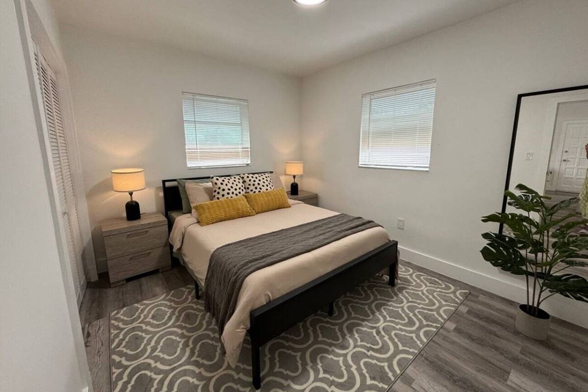 A bedroom with a bed, nightstands, lamps, a patterned rug, and a potted plant.