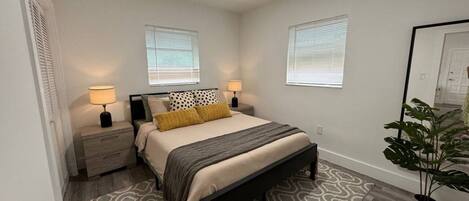 A bedroom with a bed, nightstands, lamps, a patterned rug, and a potted plant.