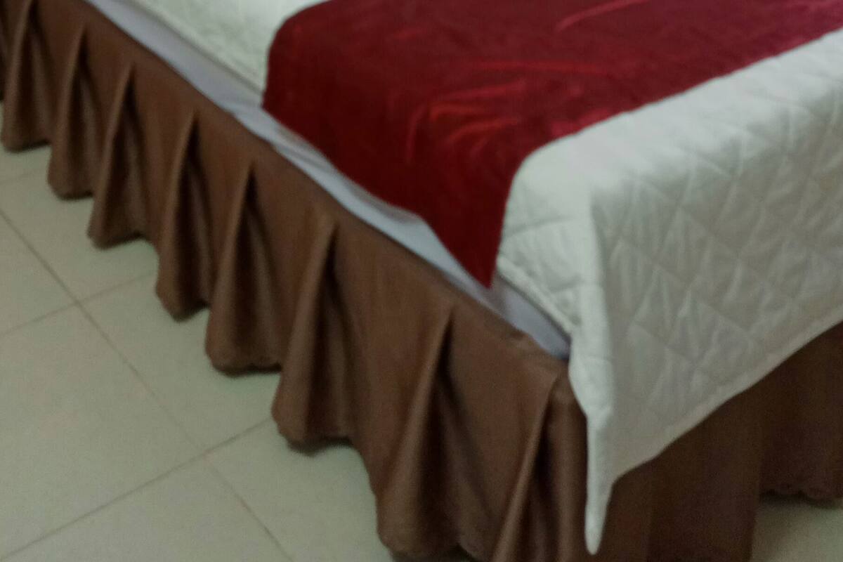 A neatly made bed with a white and red bedspread, a brown skirt, and a wall-mounted fan.