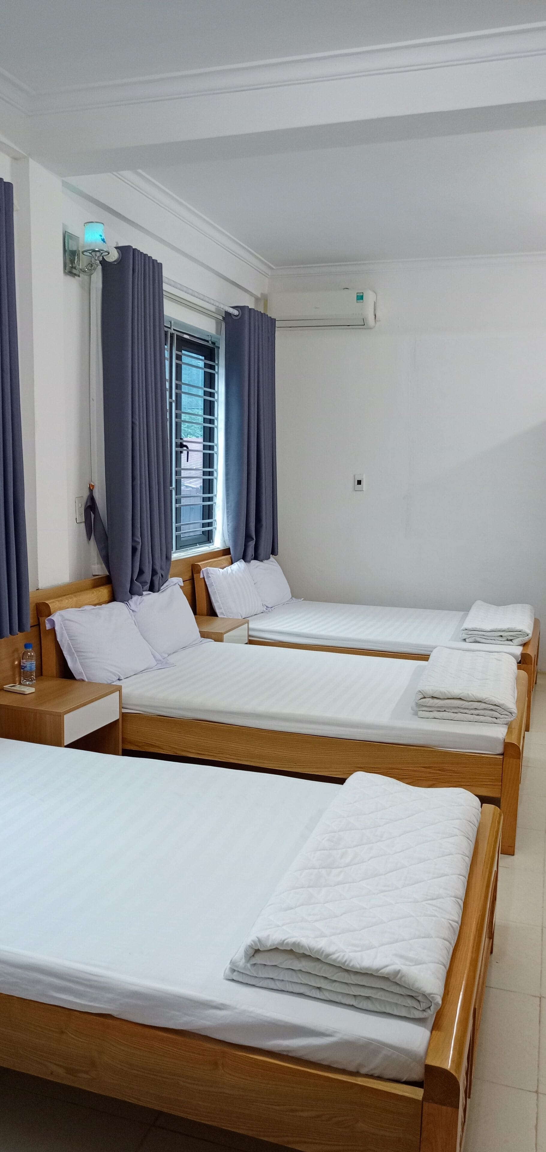 A hotel room with four single beds, a wooden headboard, a window with curtains, an air conditioning unit, and a bedside table with a bottle.