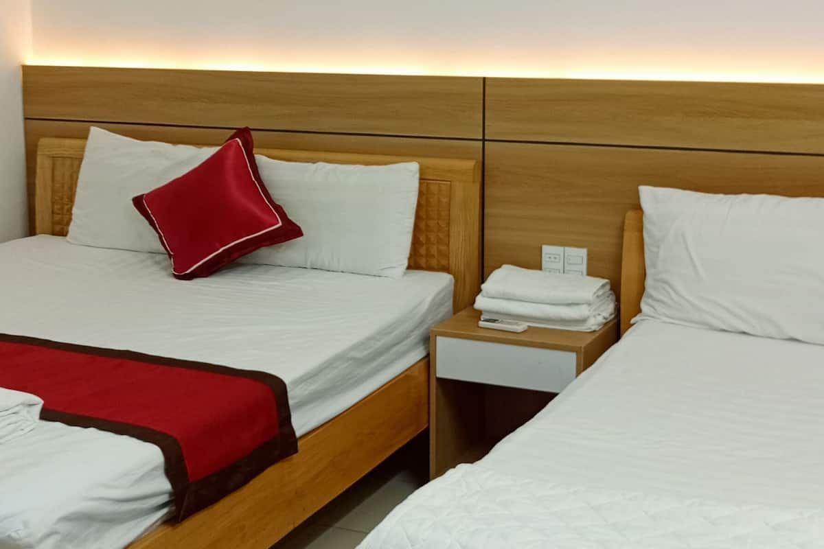 A hotel room with two wooden beds, white bedding, a red pillow, a nightstand with a lamp, and a wall-mounted light fixture.