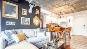 A modern living room with a grey sofa, a coffee table, and a rustic trunk. There is a kitchen area with a dining table and chairs in the background. - Rustique, NEW in Quebec city - 2 rooms + 2 bathroom (pool on roof during summer) (Québec)