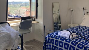 Een slaapkamer met een groot raam, een bed met een gebloemd deken, een bureau, een stoel en een hanglamp.