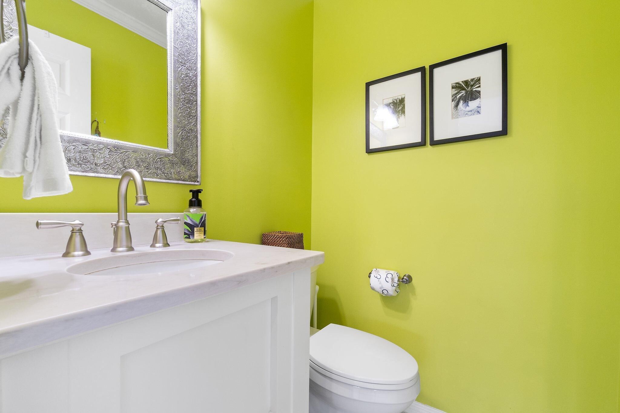 A bathroom with a white sink, three faucets, a large mirror, and a toilet. There are two framed pictures on the wall and a towel hanging on the sink.