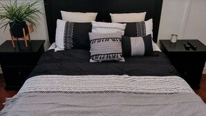 A neatly made bed with a black headboard, multiple pillows, and a grey blanket. There are two bedside tables with lamps and a potted plant.